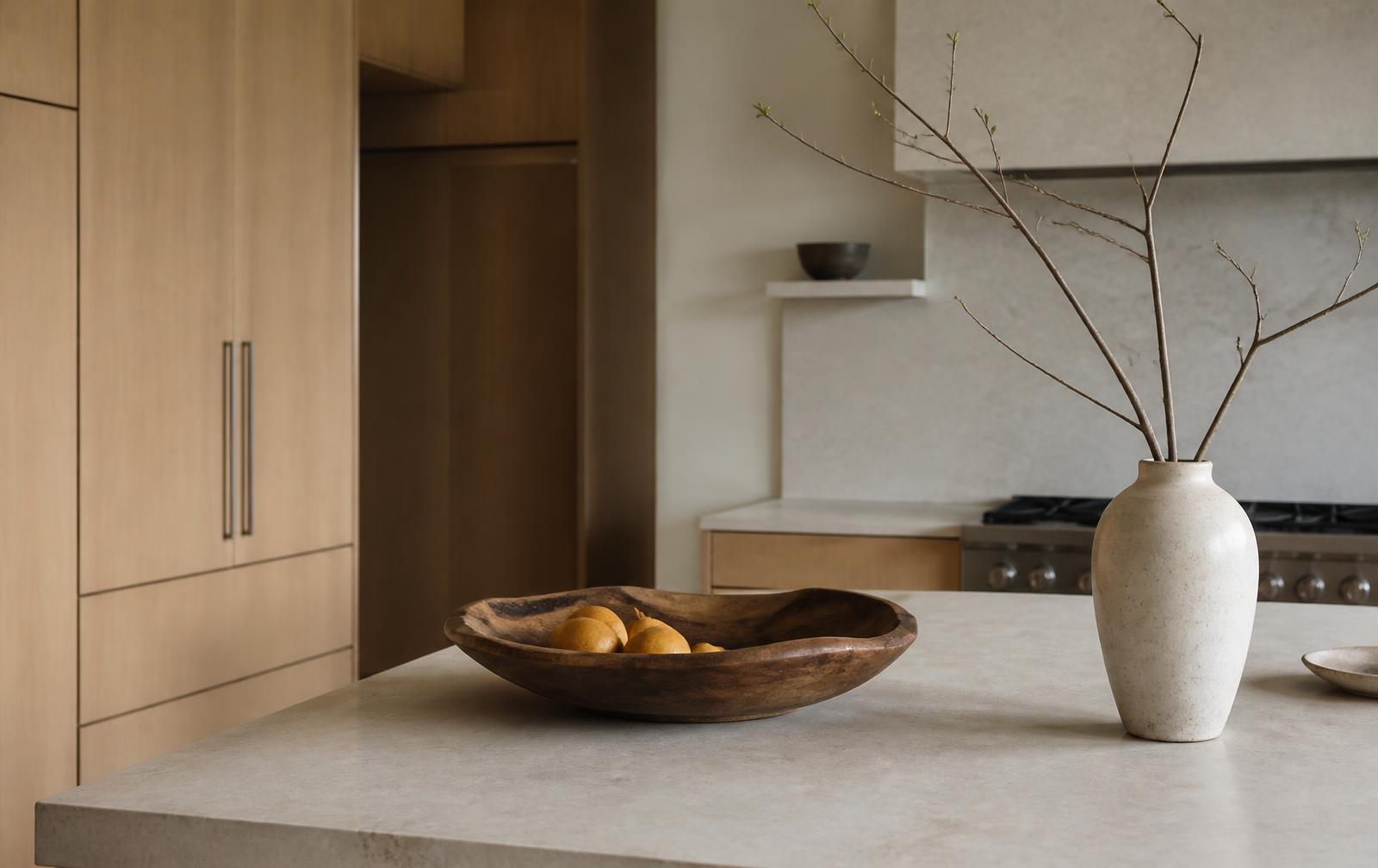 Japandi kitchen island with a large sculptural wooden bowl and a tall ceramic vase of bare branches on pale stone