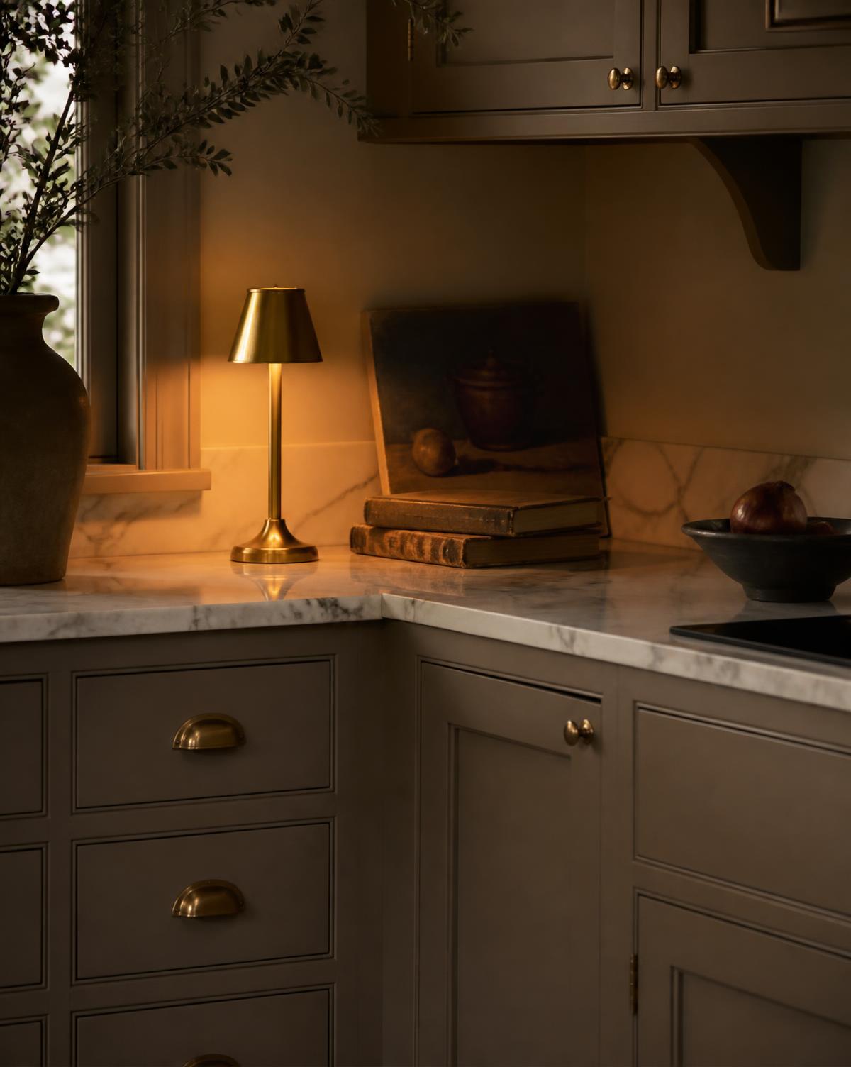 Mushroom taupe kitchen corner lit by a small brass lamp beside a leaning antique painting and old books