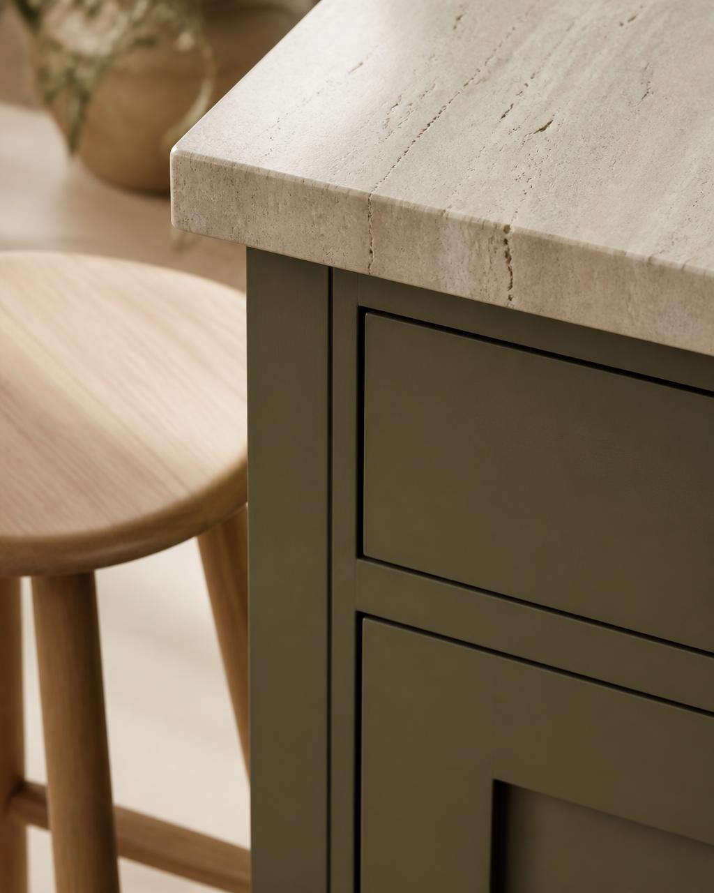 Olive green cabinetry meeting a pale travertine countertop beside a light oak stool