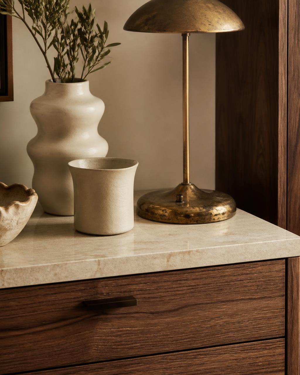 Cream ceramics and an aged brass lamp on pale stone above walnut drawers